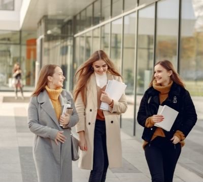 Three young women in coats, holding notebook and walking outdoors next to wall of windows