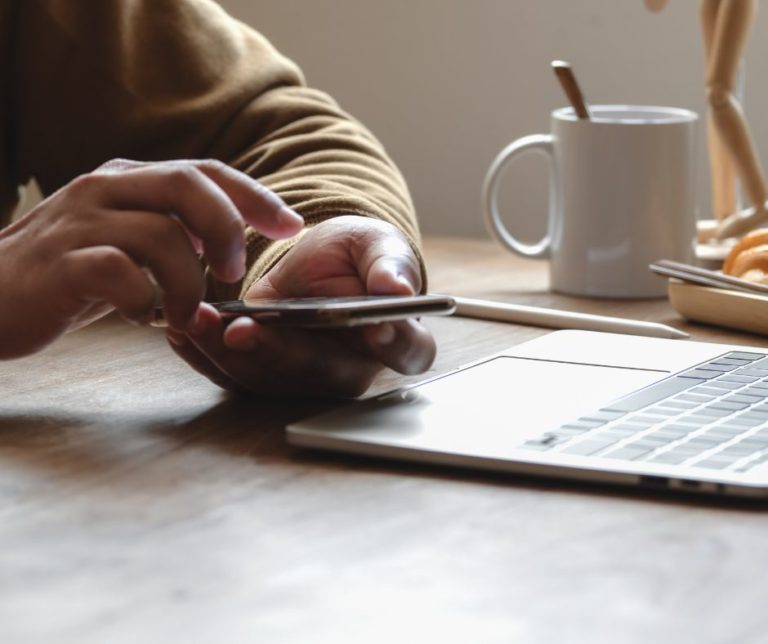 Man's hand holding phone next to mug and laptop