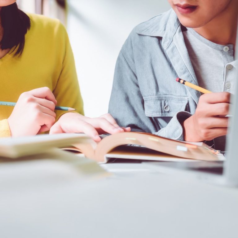 extreme close-up of two students' shoulders, arms, hands holding pencils as they look at a textbook together