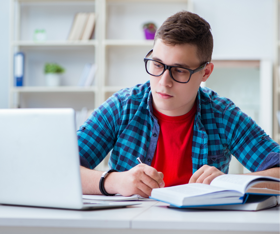 male presenting high school student with glasses, looking at open laptop and writing in notebook
